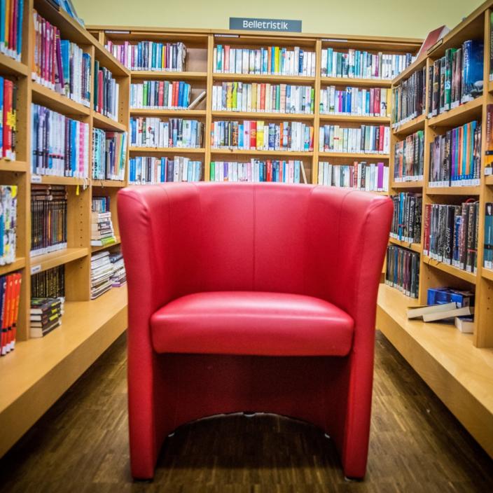 Eine Bücherei von innen, Regale mit Büchern auf drei Seiten, in der Mitte ein roter Sessel