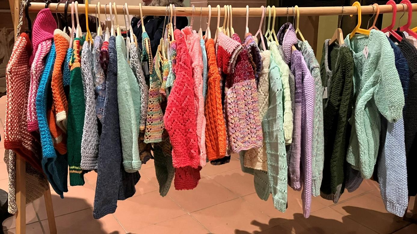 Verschiedene gestrickte und gehäkelte Kleidungsstücke hängen auf Kleiderbügeln auf einer hölzernen Stange.