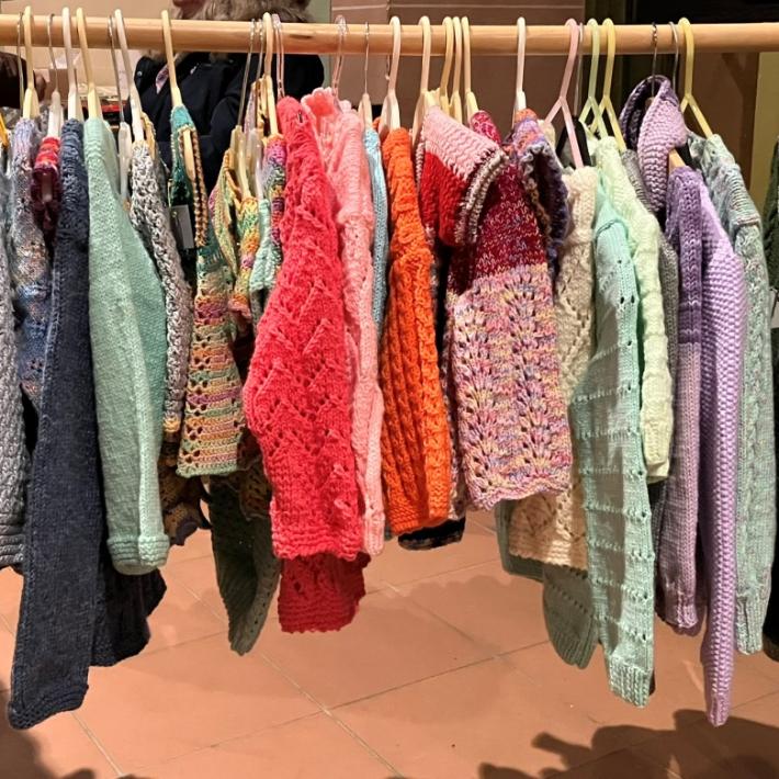 Verschiedene gestrickte und gehäkelte Kleidungsstücke hängen auf Kleiderbügeln auf einer hölzernen Stange.