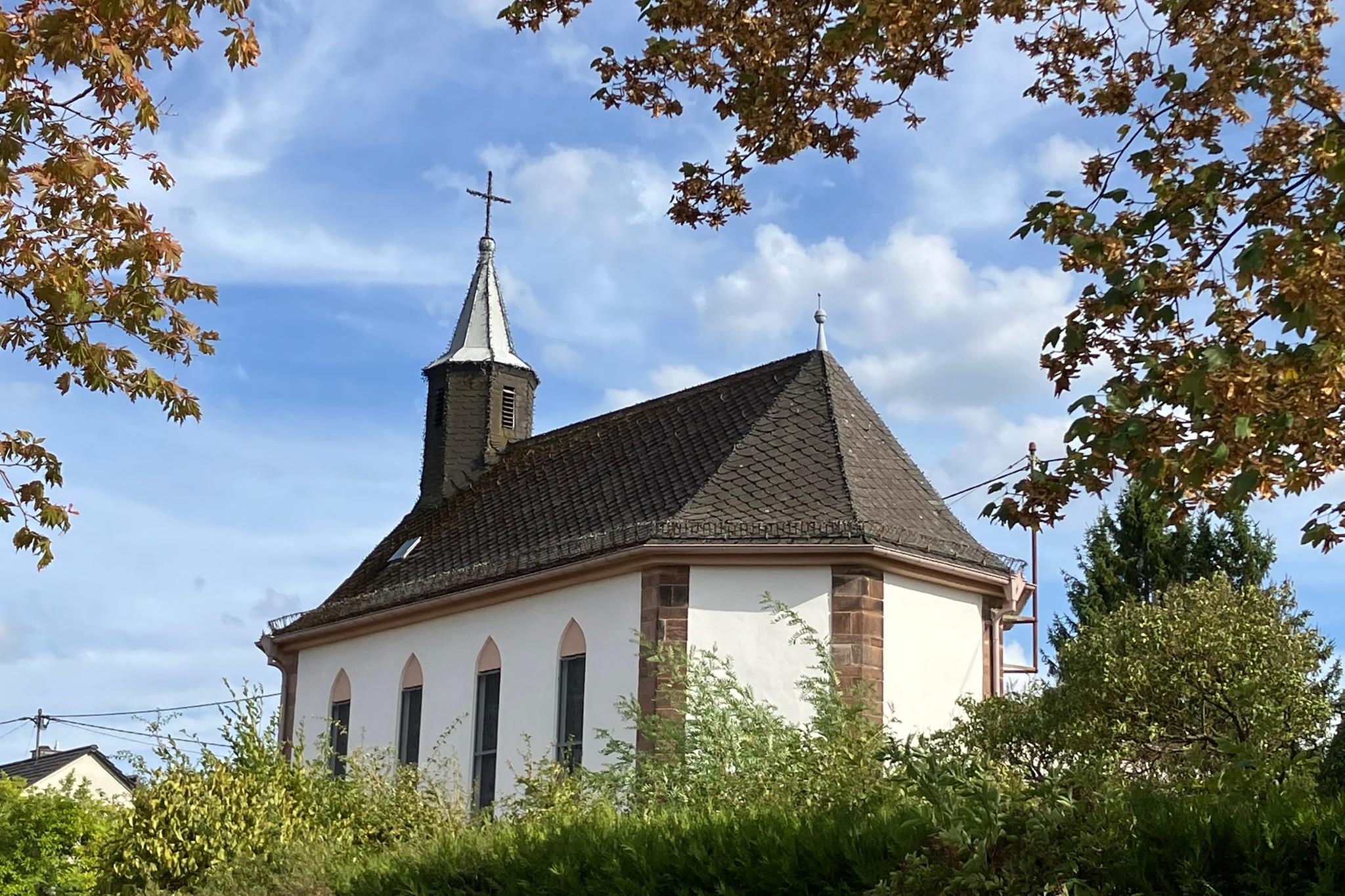 Eine kleine weiße Kapelle, aufgenommen von hinten links, bei schönem Wetter. Im Vordergrund am oberen Bildrand sieht man die Äste und Blätter von einem Baum.