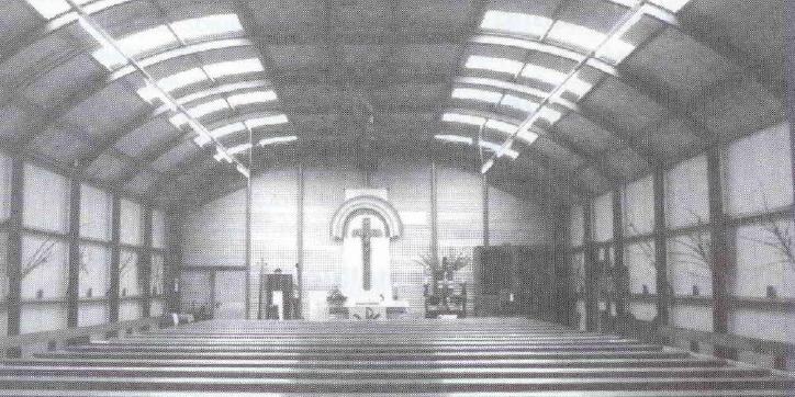 Schwarz-weiß-Bild von der Notkirche: Halle mit Dachgewölbe mit Fenstern. Der Blick geht nach vorne zum Altar. Hinter dem Altar an der Wand hängt ein großes Kreuz. Im Vordergrund sieht man die Reihen mit den Kirchenbänken.