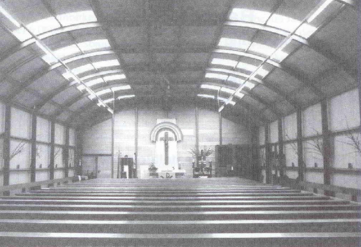 Schwarz-weiß-Bild von der Notkirche: Halle mit Dachgewölbe mit Fenstern. Der Blick geht nach vorne zum Altar. Hinter dem Altar an der Wand hängt ein großes Kreuz. Im Vordergrund sieht man die Reihen mit den Kirchenbänken.