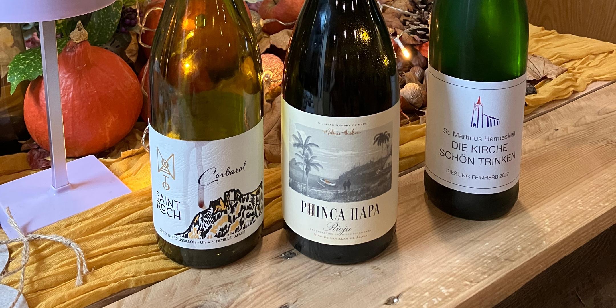 Weine mit Namen von Heiligen Drei Flaschen mit verschiedenen Weinen auf einem Holztisch. Die Weine tragen die Namen von Heiligen. Im Hintergrund: Herbstliche Dekoration.