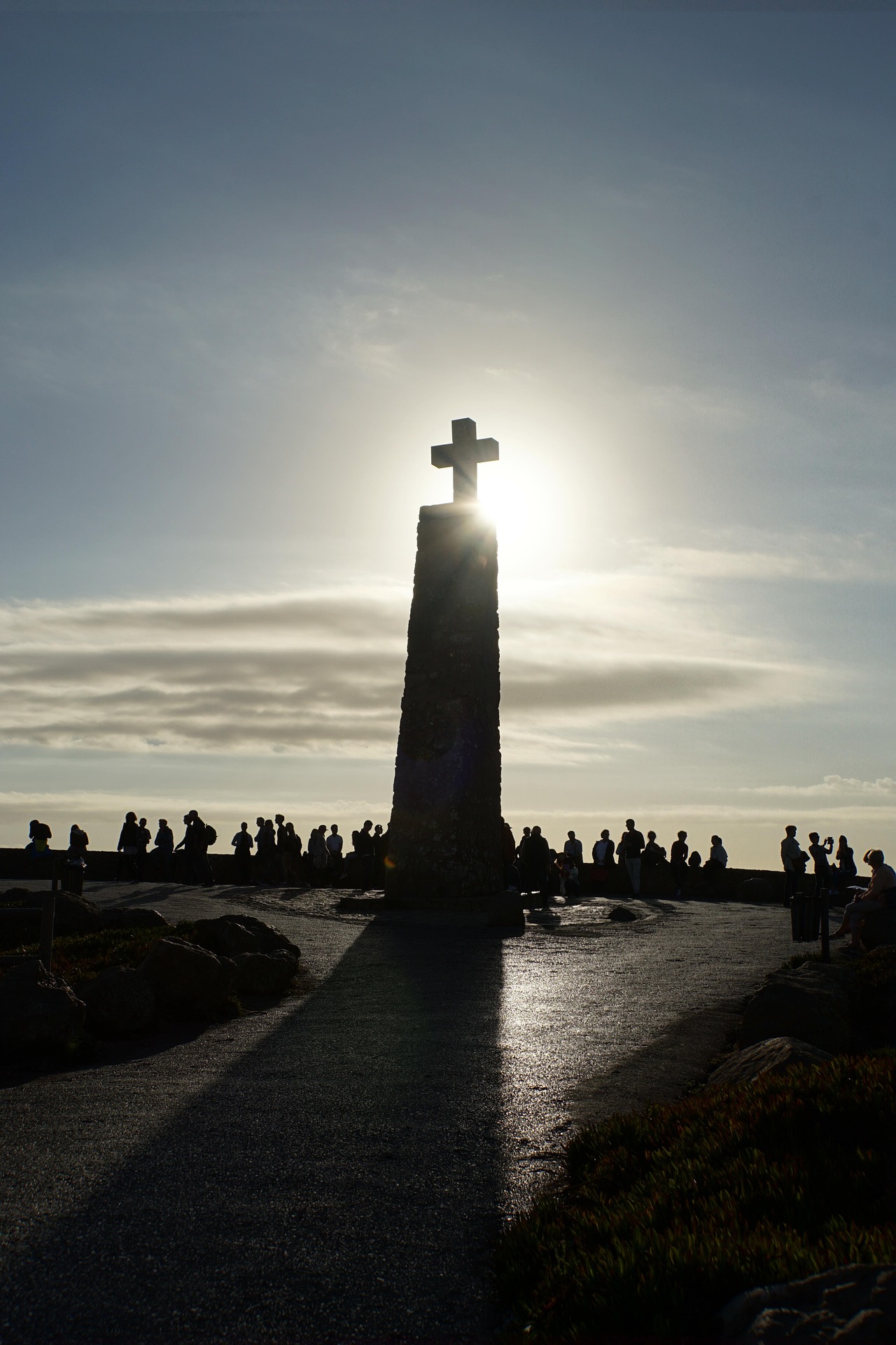 Gegenlichtaufnahme von einem Kreuz auf einem Turm. Am Fuße des Tums eine Menschenmenge