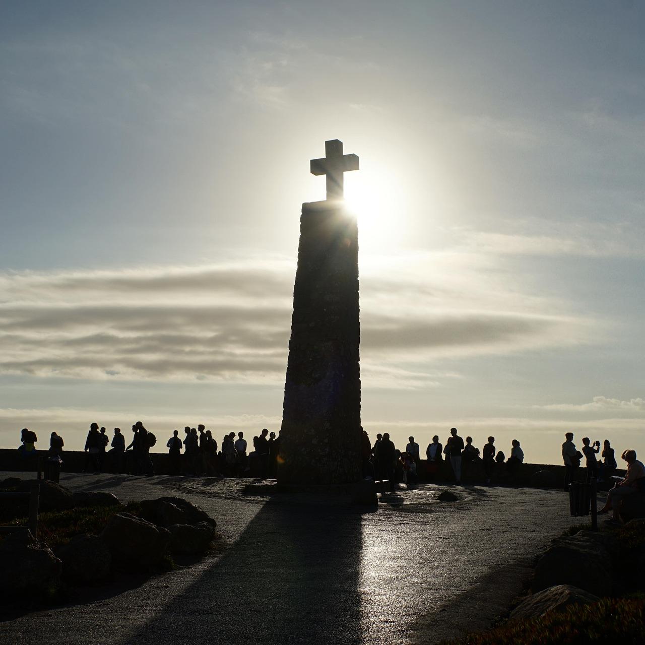 Gegenlichtaufnahme von einem Kreuz auf einem Turm. Am Fuße des Tums eine Menschenmenge